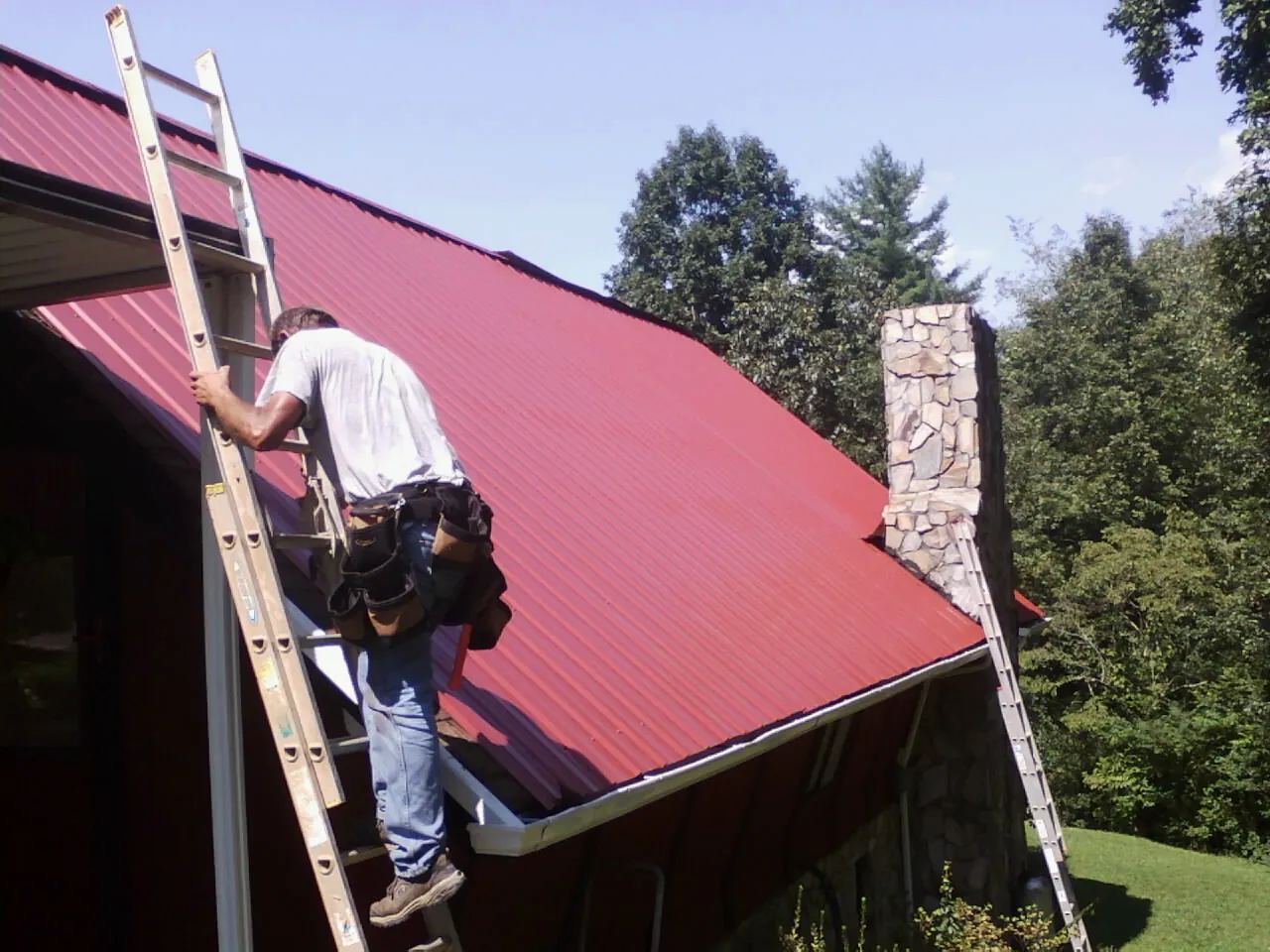 Roofer on a ladder beside a red metal roof and stone chimney — Soffit & Fascia Repair by Roofing Brothers in Rhinecliff